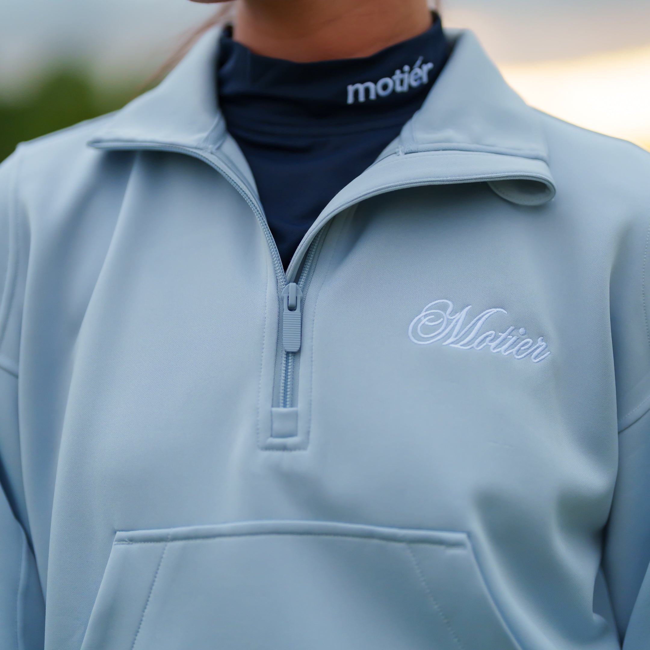 Sky blue Motier Lafayette Grand Script Scuba quarter zip sweater with a standing collar and logo embroidery, displayed on a hanger.