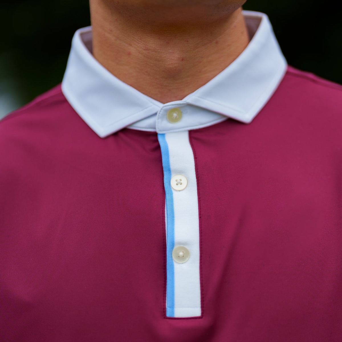 Close-up of a polo shirt collar and placket on a model. The shirt is a deep magenta color with a contrasting white collar and a white placket featuring a thin light blue stripe between the buttons.